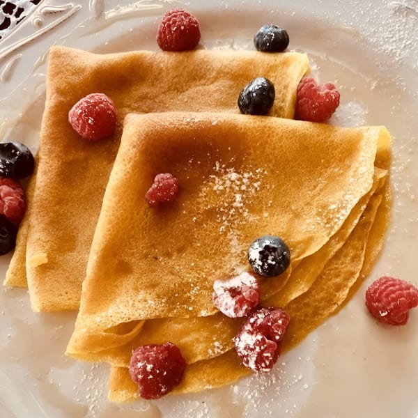 Golden crepes topped with fresh raspberries, blueberries, and powdered sugar on a white plate.