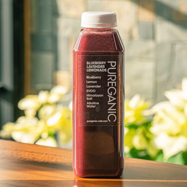 A bottle of Pureganic Blueberry Lavender Lemonade sits on a wooden surface with blurred greenery in the background.