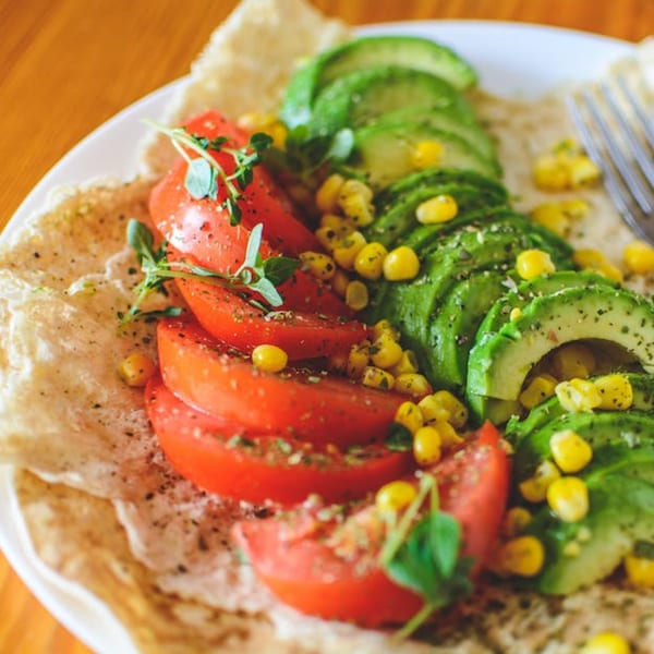 A healthy Pureganic Cafe dish with sliced tomatoes, avocado, corn, and herbs on flatbread.