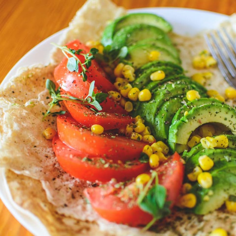 A healthy Pureganic Cafe dish with sliced tomatoes, avocado, corn, and herbs on flatbread.