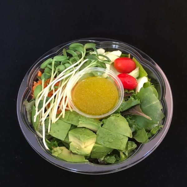 A healthy Pureganic Cafe salad with avocado, tomatoes, sprouts, and dressing in a clear container.